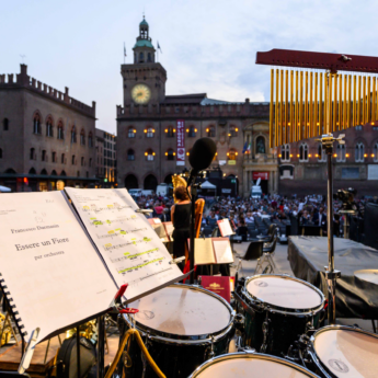 This image expresses the musical culture of Bologna, Italy. The view is from a stage of musical instruments. A musical score is visible as well as drumset. In the distance we see a singer before a live audience in a historic square.