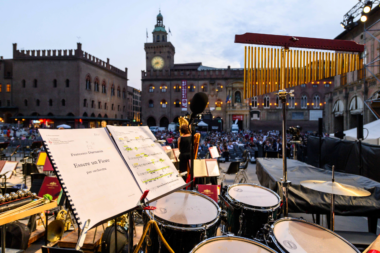 This image expresses the musical culture of Bologna, Italy. The view is from a stage of musical instruments. A musical score is visible as well as drumset. In the distance we see a singer before a live audience in a historic square.